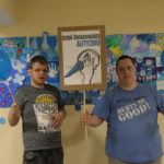 Podopieczni z tabliczka z napisem "Dzień Świadomości Autyzmu pozujący na tle obrazu stworzonego z niebieskich puzzzli.