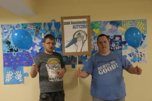 Podopieczni z tabliczka z napisem "Dzień Świadomości Autyzmu pozujący na tle obrazu stworzonego z niebieskich puzzzli.