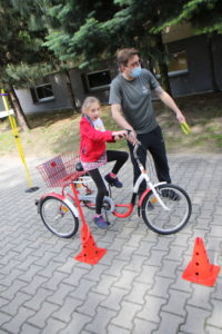 Przedszkolak pokonujący tor przeszkód na trójkowłowym rowerze z asekuracją nauczyciela.