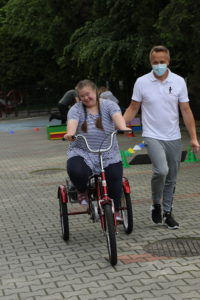 Uczennica jadąca na trójkołowym rowerze z asystą nauczyciela.