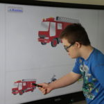 Uczeń podczas pracy dopasowywania identycznego zdjęcia pojazdu straży pożarnej na tablicy interaktywnej.