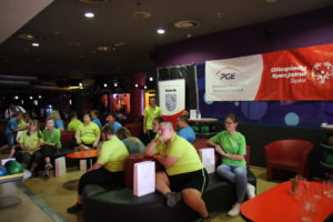 Uczestnicy turnieju bowlingowego siedzący przy stolikach na poszczególnych torach bowlingowych. W tle Banery z logortypami Olimpiad Specjalnych Miasta Rybika oraz PGE.