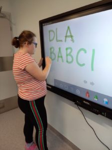 Uczennica pisząca po śladzie napis "dla babci" na tablicy interaktywnej.