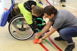 Uczeń poruszający się na wózku podczas aktywności nakładania piłeczki z pomocą trenera.