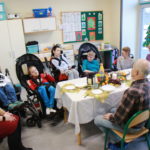 Podopieczni na wózkach rehabilitacyjcnych wraz z wychowawcami przy stole podczas wigilijki.