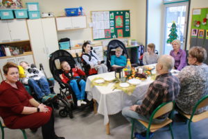Podopieczni na wózkach rehabilitacyjcnych wraz z wychowawcami przy stole podczas wigilijki.