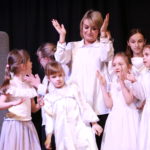 Występujące w białych sukniach przedszkolaki, uczniowie wraz z nauczycielką pokazujący gesty piosenki.