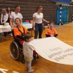 Uczniowei na wózkach oraz wolontariusze trzymający flagę Olimpiad Specjalnych