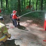 Ucznieiwe oraz strażacypodczas poazowej aktywności lania wodą z węża strażackeiego do celu.